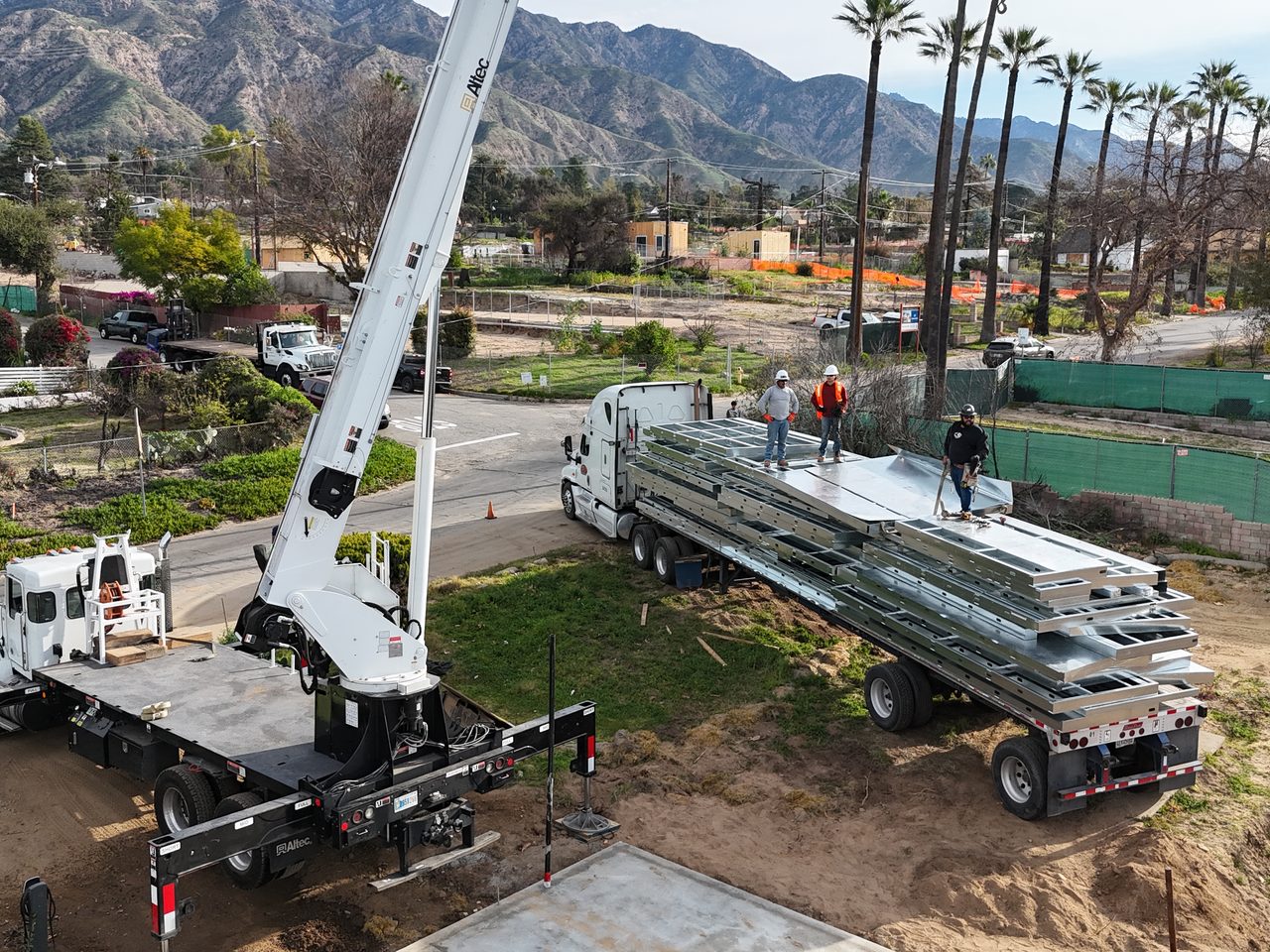 A boom crane and flatbed delivering panelized light gauge steel to a foothill jobsite, with the San Gabriel mountains behind