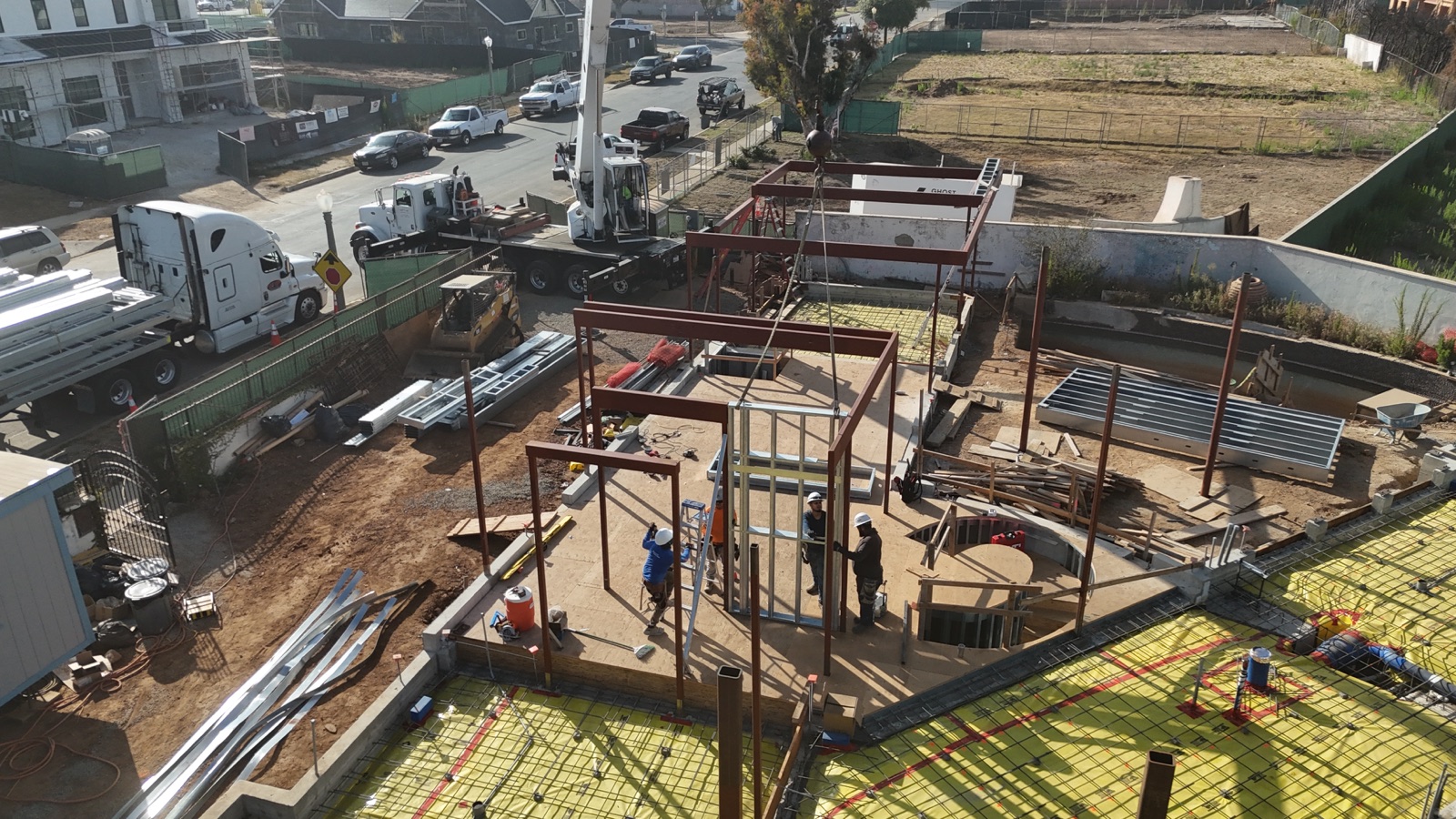 Aerial of the red-oxide structural steel frame going up at 777 Ocampo, a crane setting a beam as ironworkers frame the level