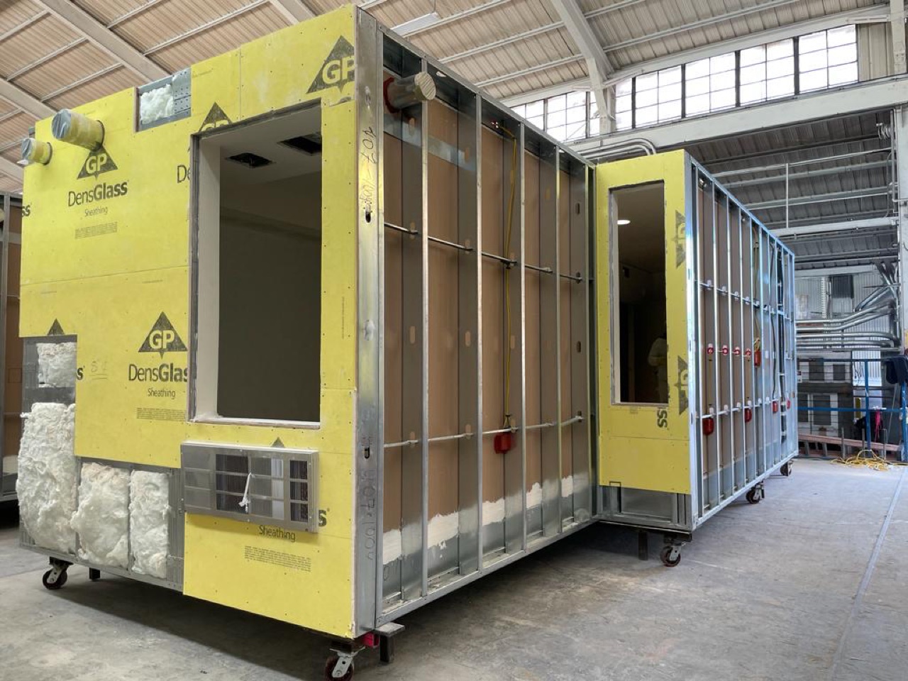 A complete steel-framed volumetric module with yellow DensGlass exterior sheathing and a framed window opening, built on the factory floor